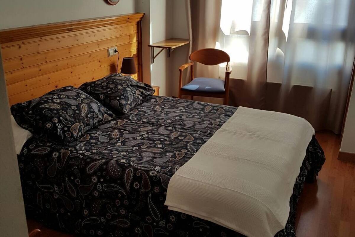 Een slaapkamer met een houten hoofdeinde, een bed met een geborduurde spreidekking, een nachtkastje met een lamp, een stoel en een raam met gordijnen.
