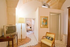 Una habitación con una televisión sobre una mesa de madera, una lámpara, una silla con cojín, un cuadro en la pared y suelo de baldosas.