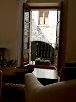 Un umbral abierto que da a una construcción de piedra con un ventanal de madera, una mesa pequeña con una cesta y una silla.