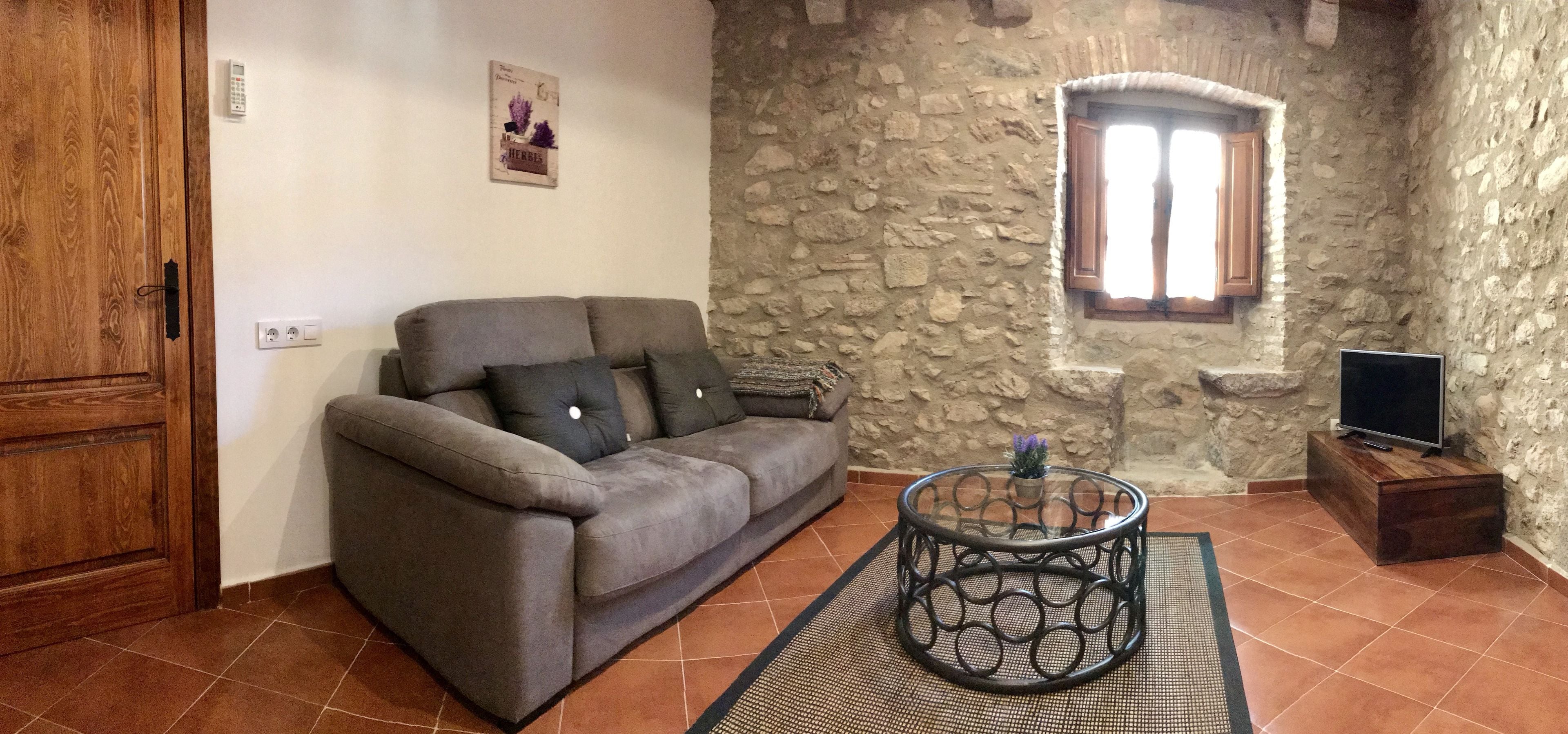 Una sala de estar con una pared de piedra, una puerta de madera, un sofá gris, una mesa de centro de vidrio y un televisor sobre un mueble de madera.