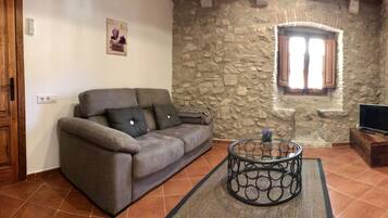Una sala de estar con una pared de piedra, una puerta de madera, un sofá gris, una mesa de centro de vidrio y un televisor sobre un mueble de madera.