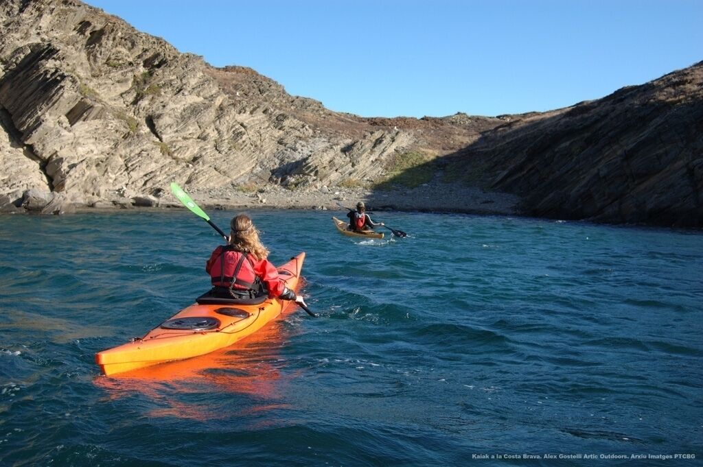 Two kayakers on a calm body of water with rocky cliffs in the background.