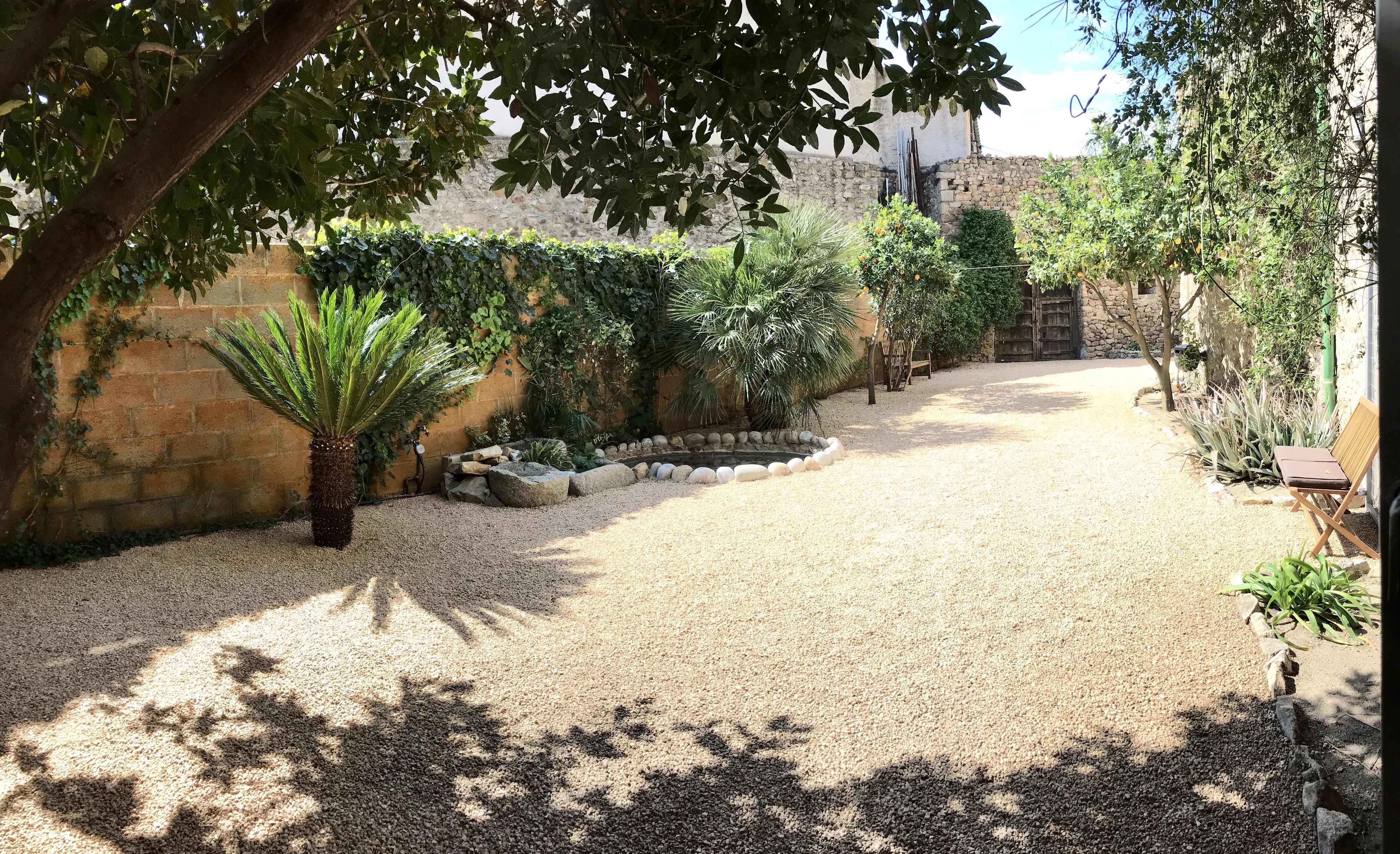 A gravel courtyard with a stone wall, a palm tree, and various shrubs.