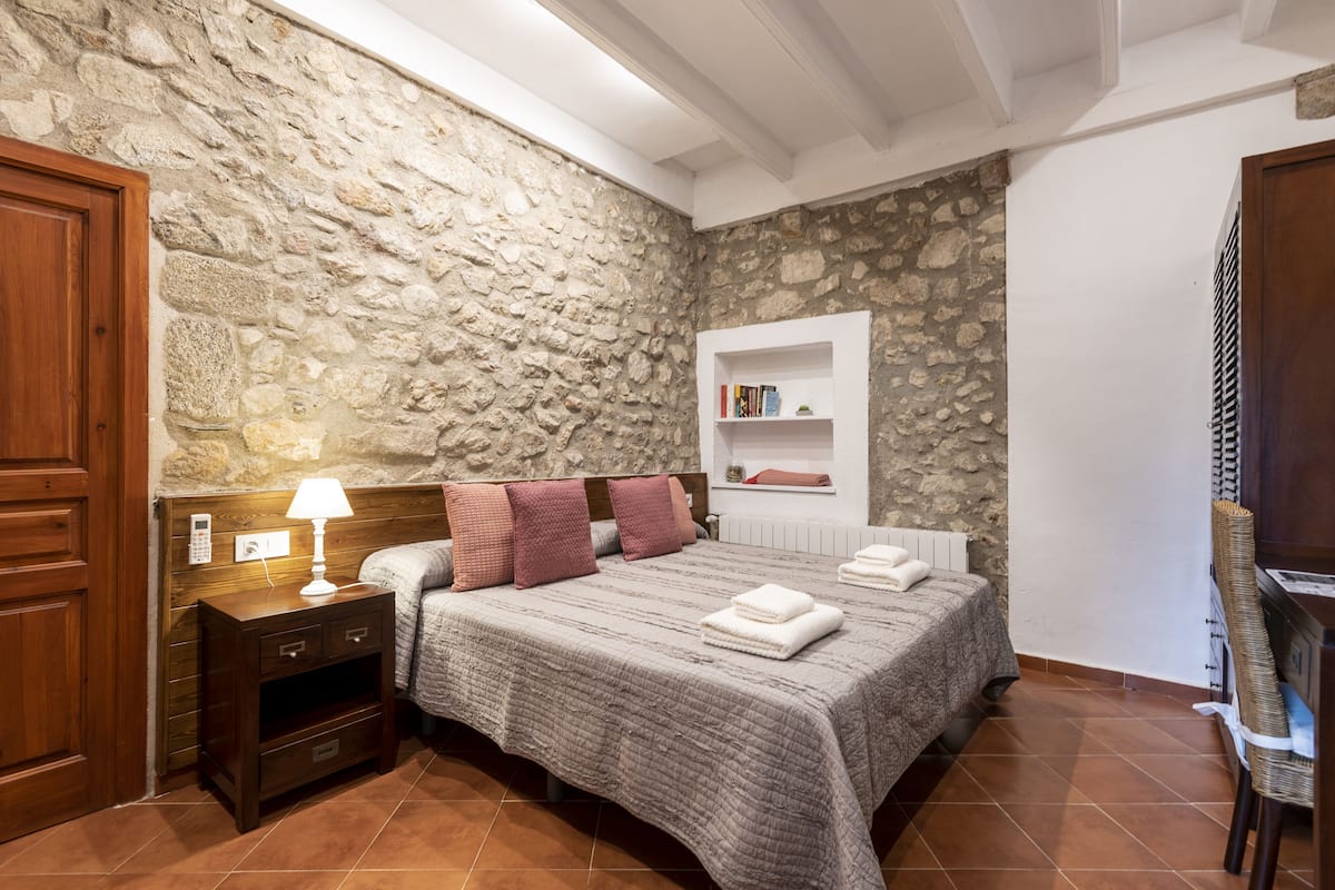 Un dormitorio con pared de piedra, cama de madera, mesita de noche con lámpara y estantería.