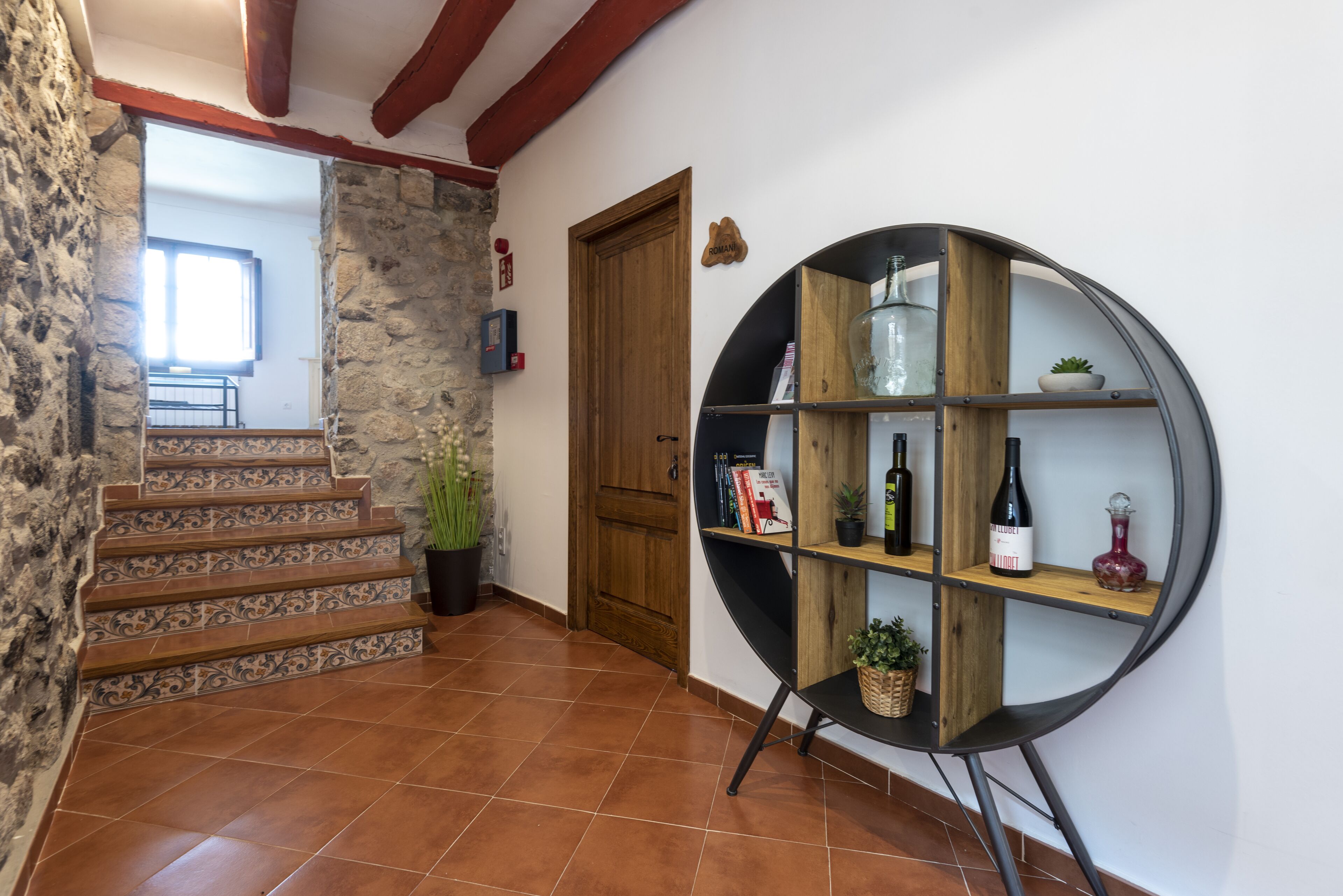 A staircase with patterned tiles leading to a room with a wooden door, a bookshelf with wine bottles, and a potted plant.