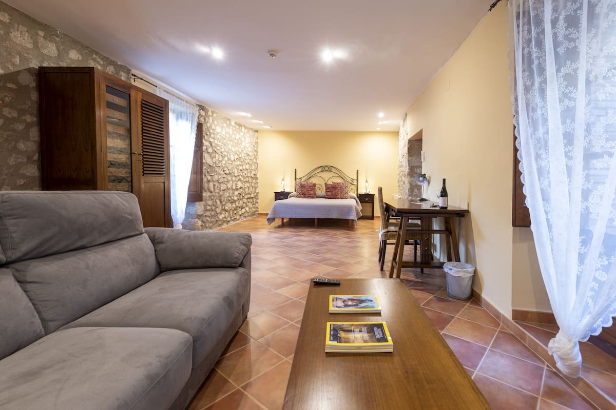 Un salón acogedor con sofá, una mesa de centro de madera con libros, una cama con cabecero y una pared de piedra.