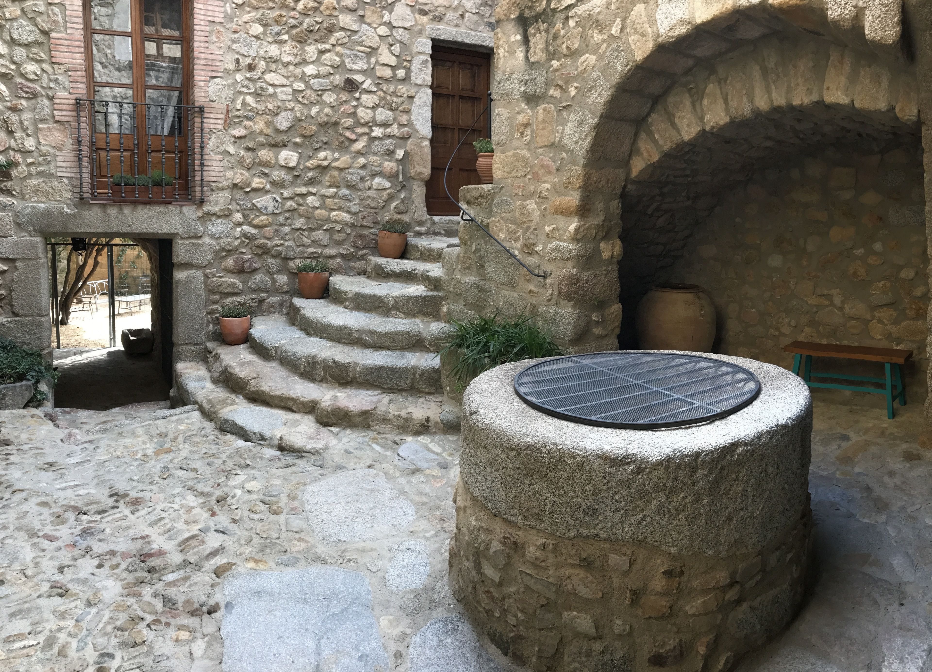 Un patio de piedra con un pozo circular, escalones que conducen a una puerta de madera y plantas en macetas.
