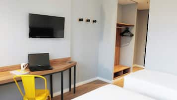 Une chambre d’hôtel moderne avec un bureau en bois, un ordinateur portable, une chaise jaune, un lit, un téléviseur fixé au mur et un dressing.