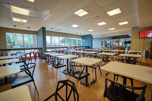 Una cafetería con varias mesas y sillas, ventanas amplias y una pantalla de televisión.