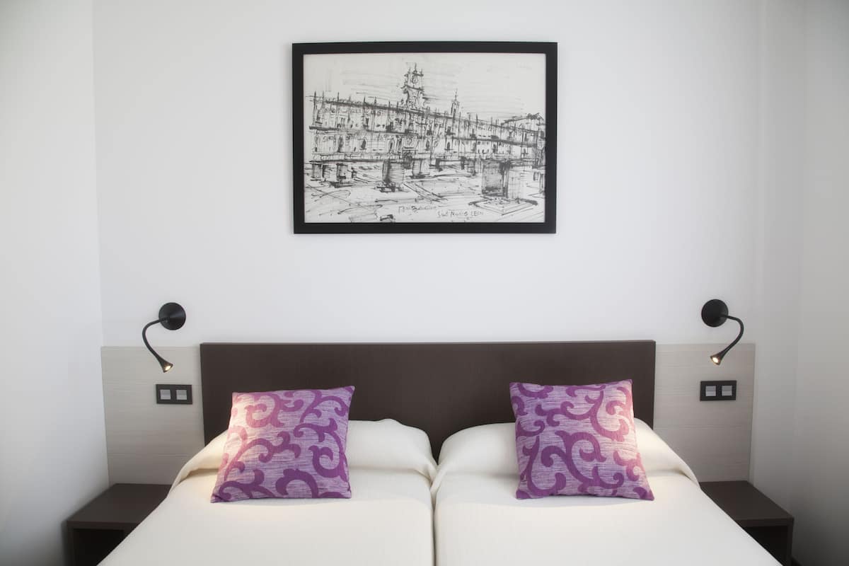 A double bed with two purple patterned pillows, a framed black and white cityscape artwork above it, and two bedside lamps on either side.