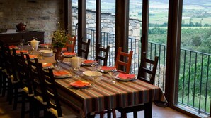 A dining area with a long table set for a meal, large windows offering a view of the countryside, and a stone wall in the background. - O Chardinet d'a Formiga (La Fueva)