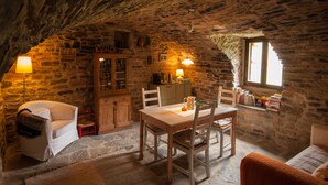 A rustic stone room with a wooden table and chairs, a small cabinet, and a window with books on a shelf. - O Chardinet d'a Formiga (La Fueva)