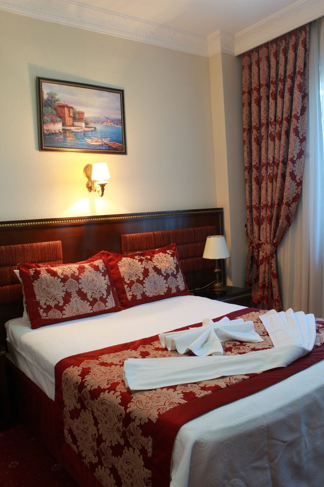 A hotel room with a large bed, a headboard, a nightstand with a lamp, a painting on the wall, and a window with curtains.