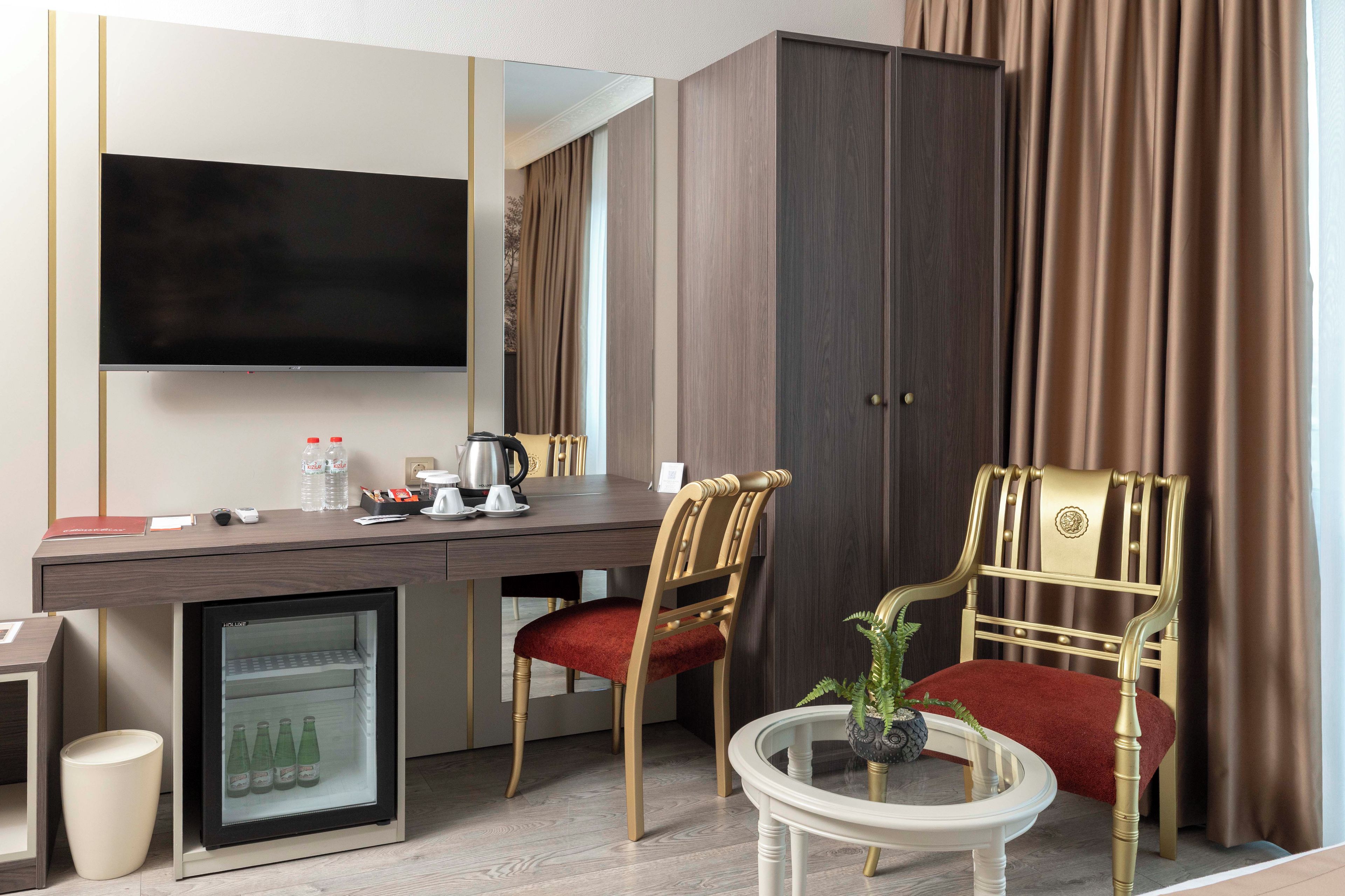 A hotel room with a desk, two chairs, a television, a minibar, and a small table with a plant.