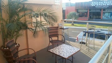 Varanda com cadeiras de vime e uma pequena mesa, com vista para uma rua com loja.