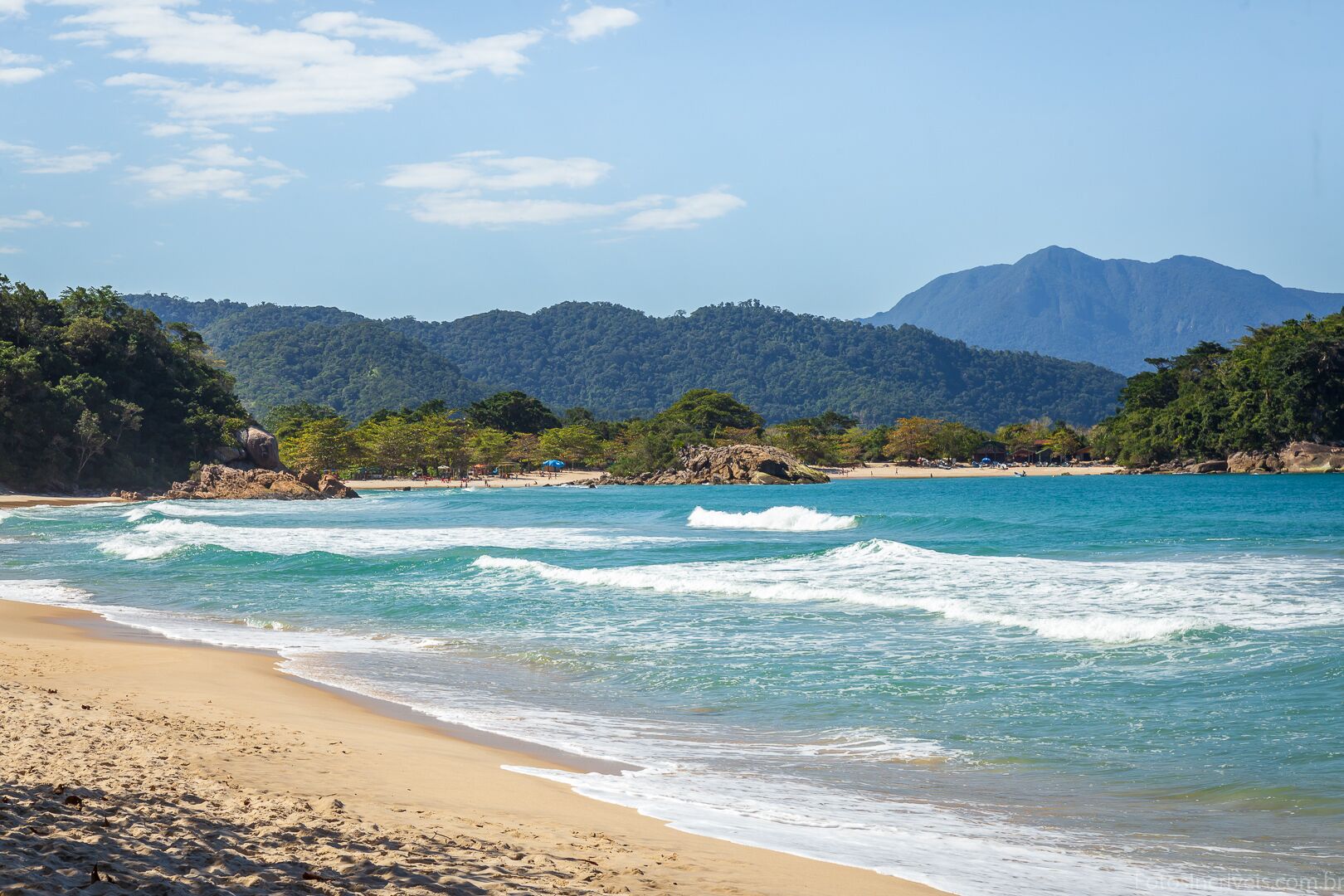 Uma praia com águas turquesa cristalinas, areal arenoso e uma montanha arborizada ao fundo.