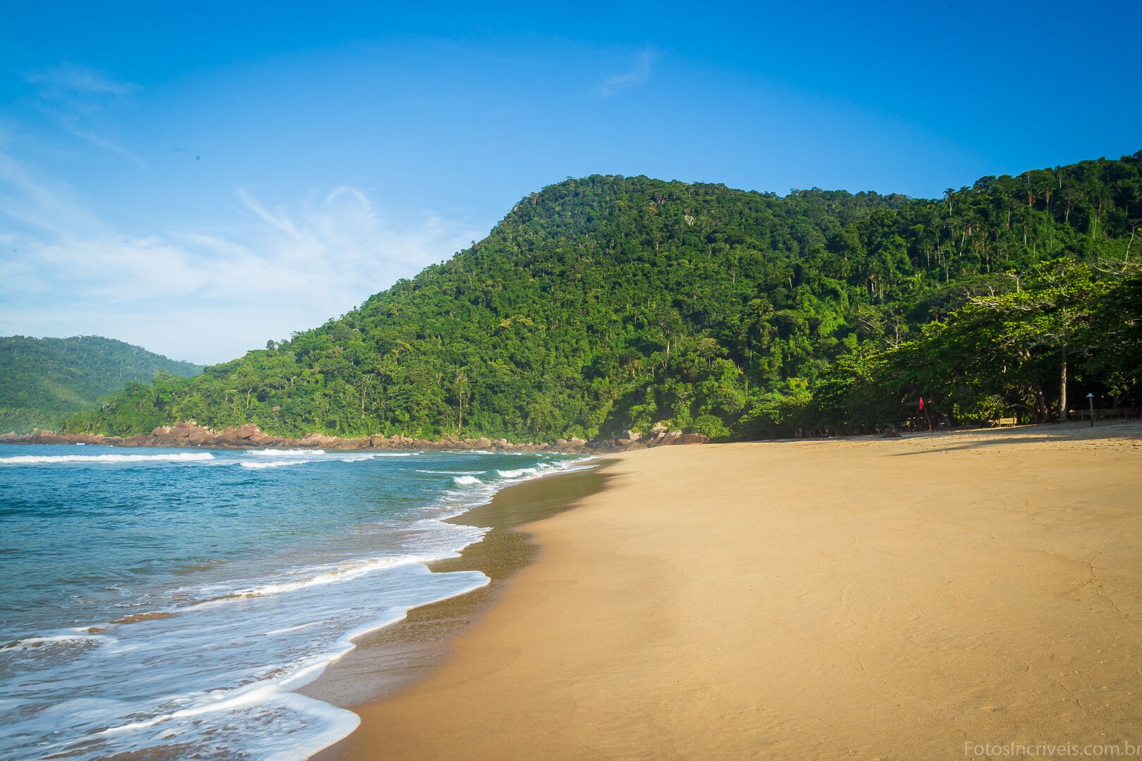 A beach with clear blue water, sandy shore, and a forested hill in the background.