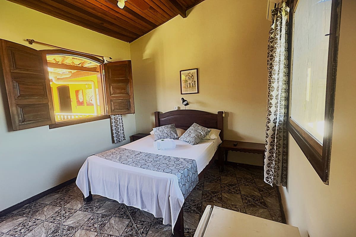 Un dormitorio con cama, cabecera de madera, mesita de noche, lámpara, ventana con persianas abiertas y una puerta.