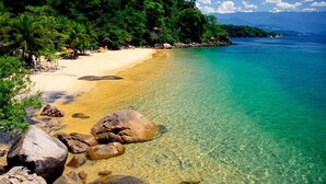 Una playa con agua turquesa cristalina, grandes rocas en primer plano y abundante vegetación verde al fondo.