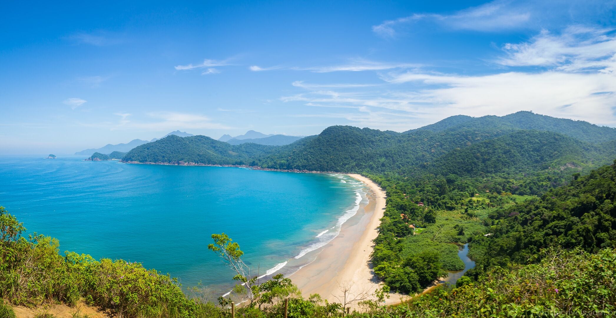 A coastal landscape with a beach, forested hills, and clear blue waters.
