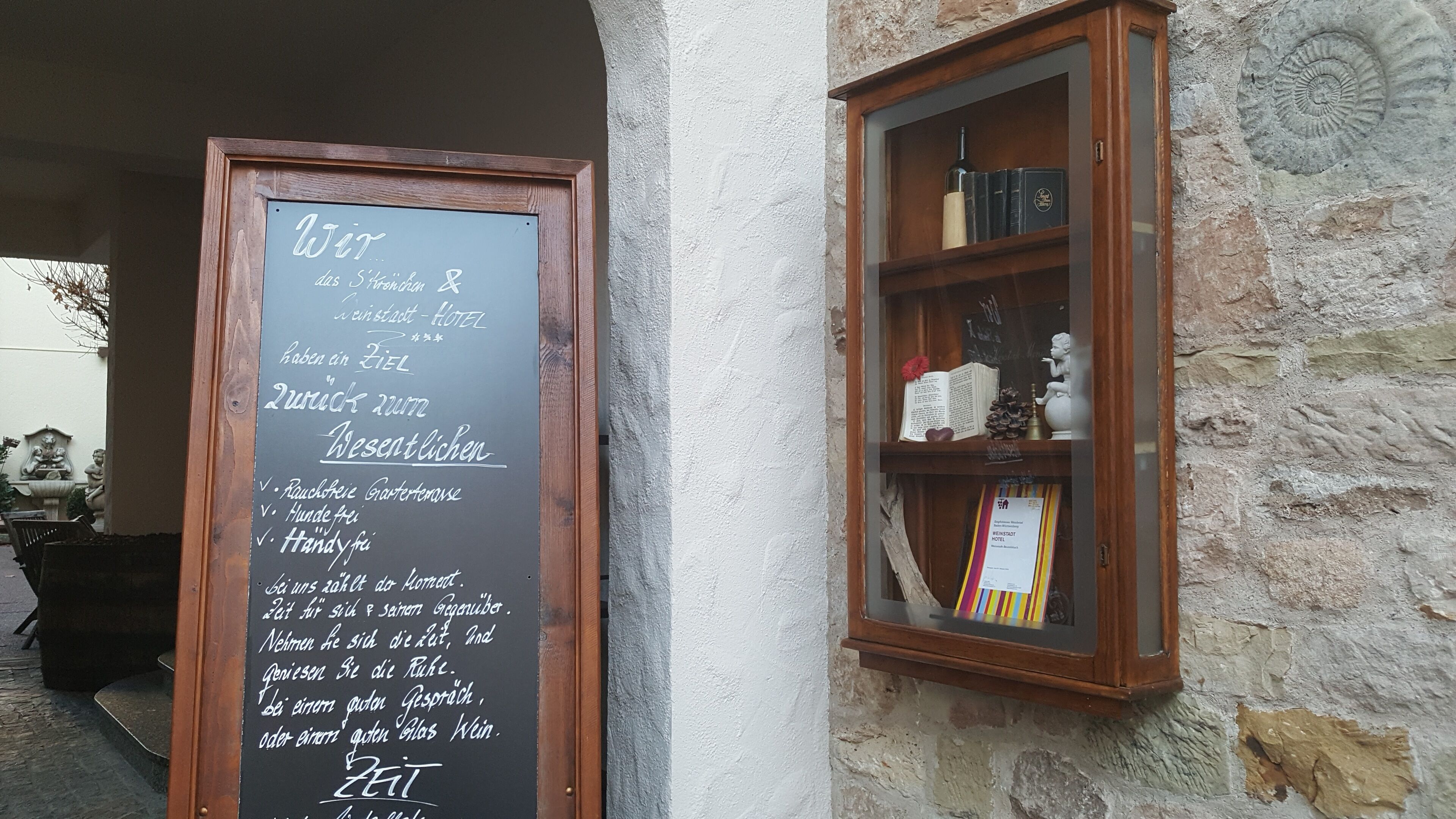 Eine Kreidetafel mit Text auf Deutsch, ein Holzregal mit Büchern und Dekoration, sowie eine Steinwand.