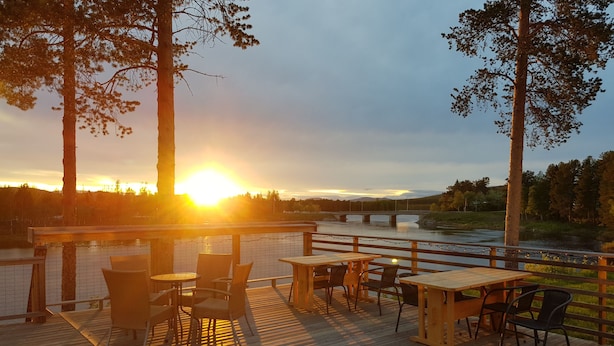 A wooden deck with tables and chairs, overlooking a calm river and a bridge. - Sorsele River Hotel (Sorsele)