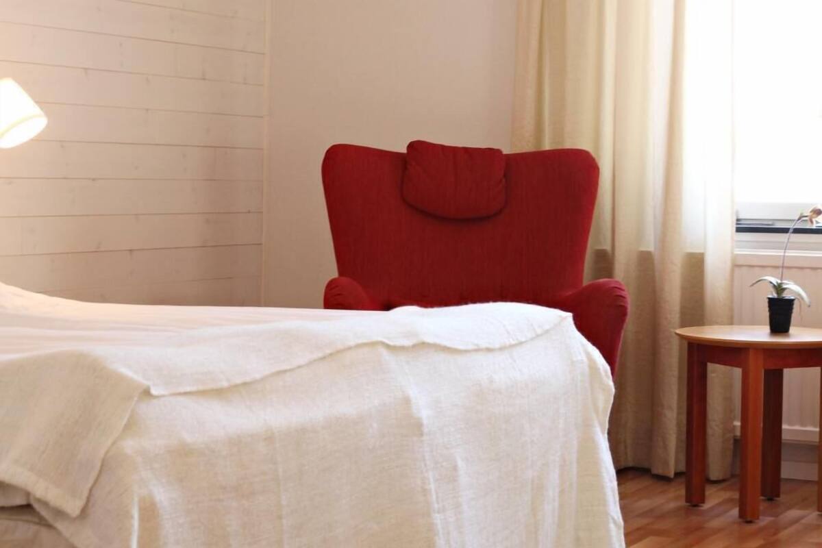 Een slaapkamer met een bed, een rode stoel, een tafeltje met een plant en een raam met gordijnen.
