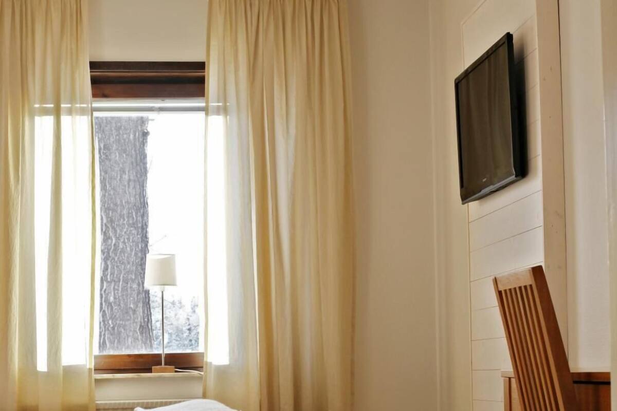 Een slaapkamer met een bed, een stoel, een aan de muur bevestigde televisie en een raam met gordijnen.