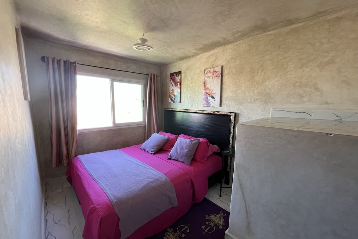A bedroom with a bed, curtains, a ceiling fan, and artwork on the wall.