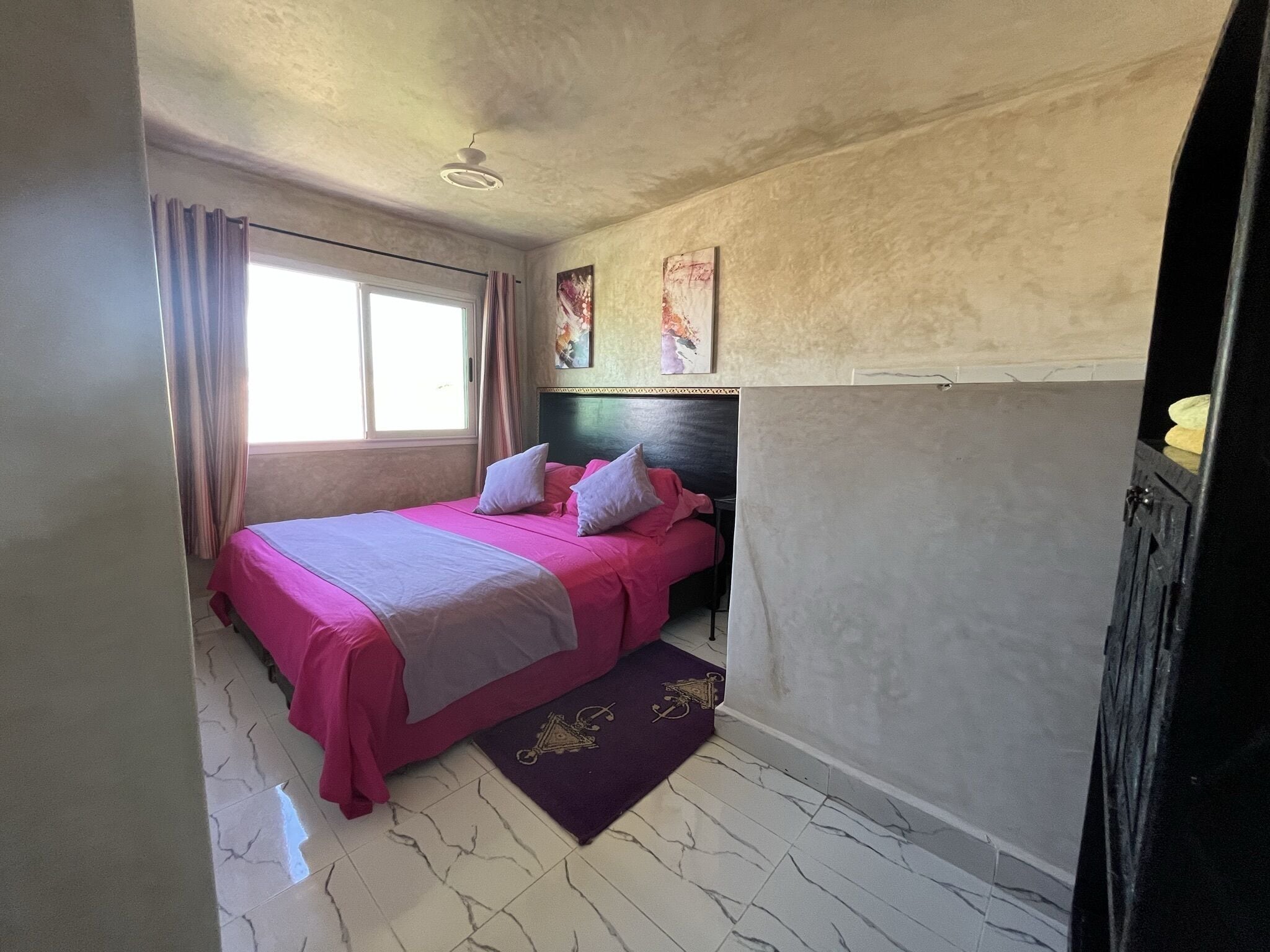 A bedroom with a bed, curtains, a ceiling fan, a window, and a wall with a painting.