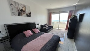 A bedroom with a large bed, a nightstand, a dresser, a TV, and a balcony with a view of the sea. - Riad Dar Naima Imsouane (Imsouane)