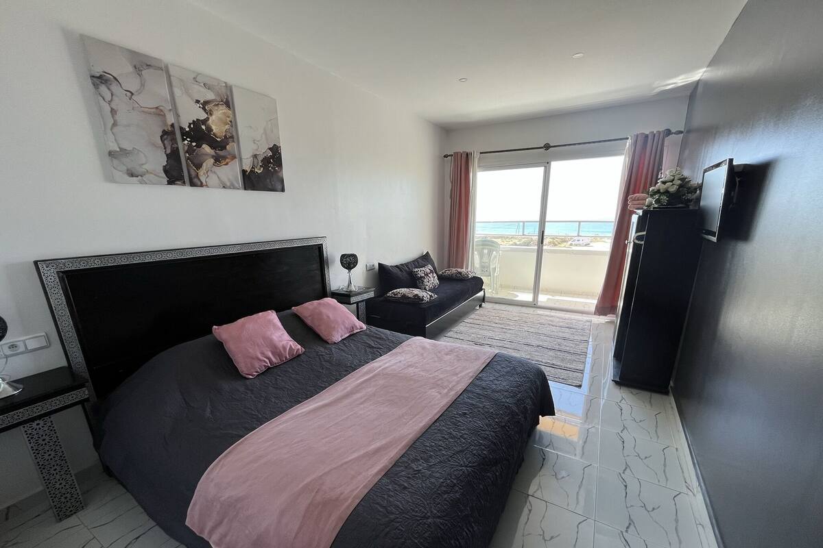 A bedroom with a large bed, a nightstand, a dresser, a TV, and a balcony with a view of the sea.