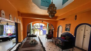 A room with a patterned floor, a sofa, a dining table with a vase of flowers, and a view of an outdoor area with a blue door and a plant. - Riad Dar Naima Imsouane (Imsouane)