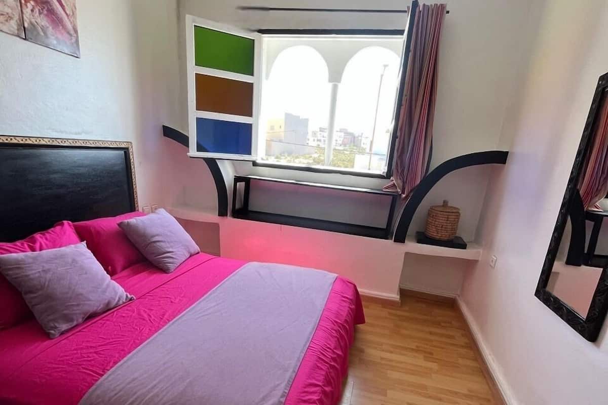 A bedroom with a bed, a window with curtains, a shelf with decorative items, and a painting on the wall.