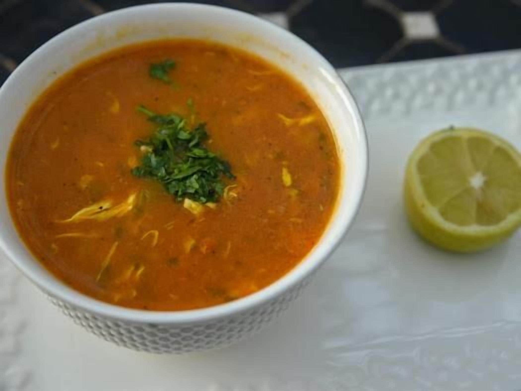 Un bol de soupe agrémenté d’une garniture, accompagné d’une demi-citron sur une surface blanche.