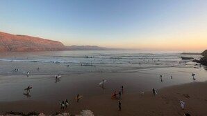 A beach scene with people walking and surfing, and a rocky cliff in the background. - Riad Dar Naima Imsouane (Imsouane)