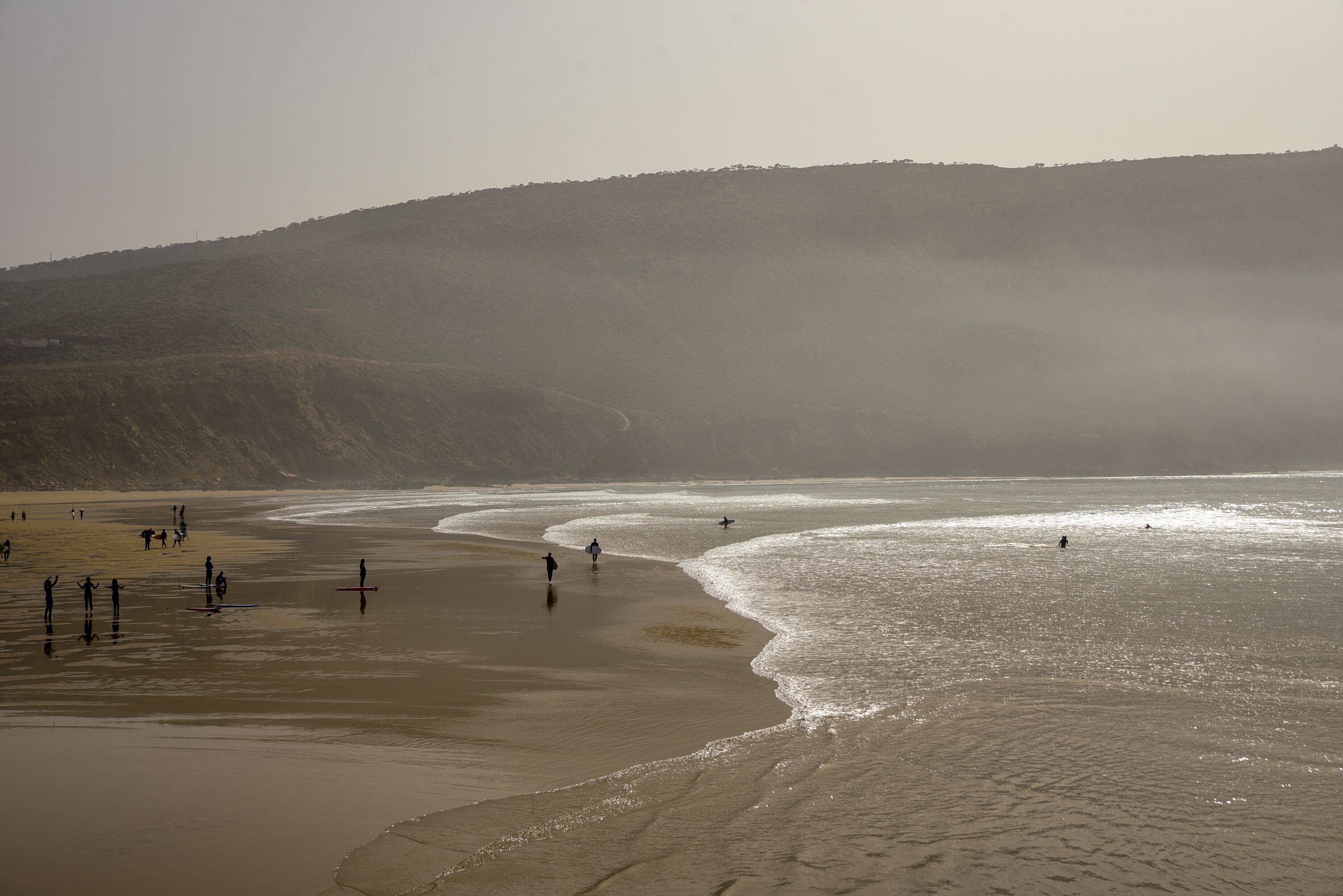 Una playa con gente caminando y practicando surf, orilla arenosa y un paisaje montañoso al fondo.