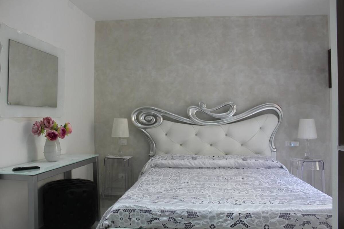 A bedroom with a tufted headboard, a white bedspread with a floral pattern, a bedside table with a lamp, and a small bedside table with a vase of flowers.