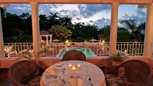Un área de comedor al aire libre con sillas de mimbre y una mesa preparada para dos, con vistas a una piscina y abundante vegetación.