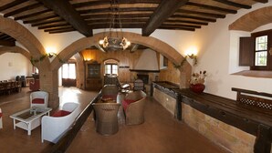 A spacious interior with high ceilings, exposed wooden beams, and arched brick walls. There are multiple seating areas with wicker furniture and a central fireplace. - Podere Agrituristico Luchiano (Amelia)