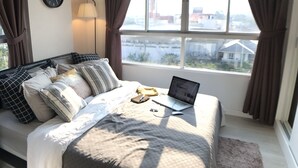 Une chambre à coucher avec un lit, un ordinateur portable, une horloge et une vue sur les bâtiments par la fenêtre.