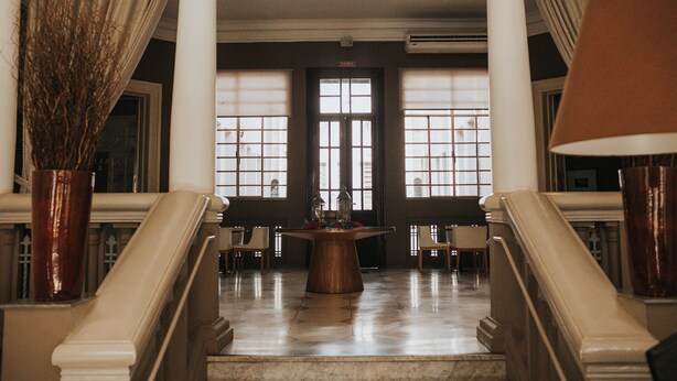 Un lobby espacioso con piso de mármol, una mesa de madera en el centro y una escalera con barandales blancos.