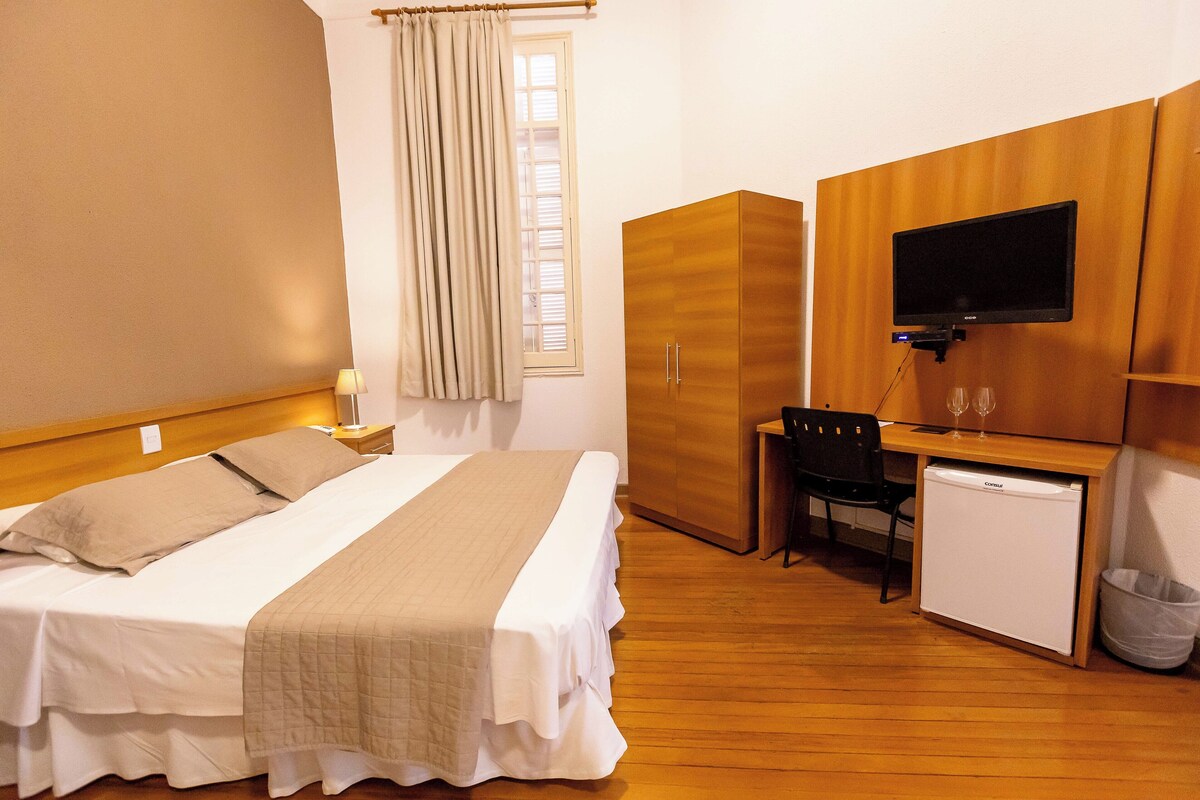 Una camera d'albergo con un letto, una scrivania con un computer, una televisione e un piccolo frigorifero.