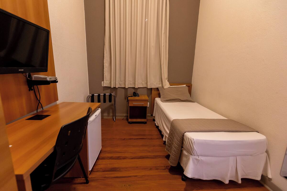A small, compact room with a wooden desk, a bed, a TV mounted on the wall, and a window with curtains.