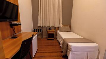 Una habitación pequeña y acogedora con escritorio de madera, cama, televisor montado en la pared y ventana con cortinas.