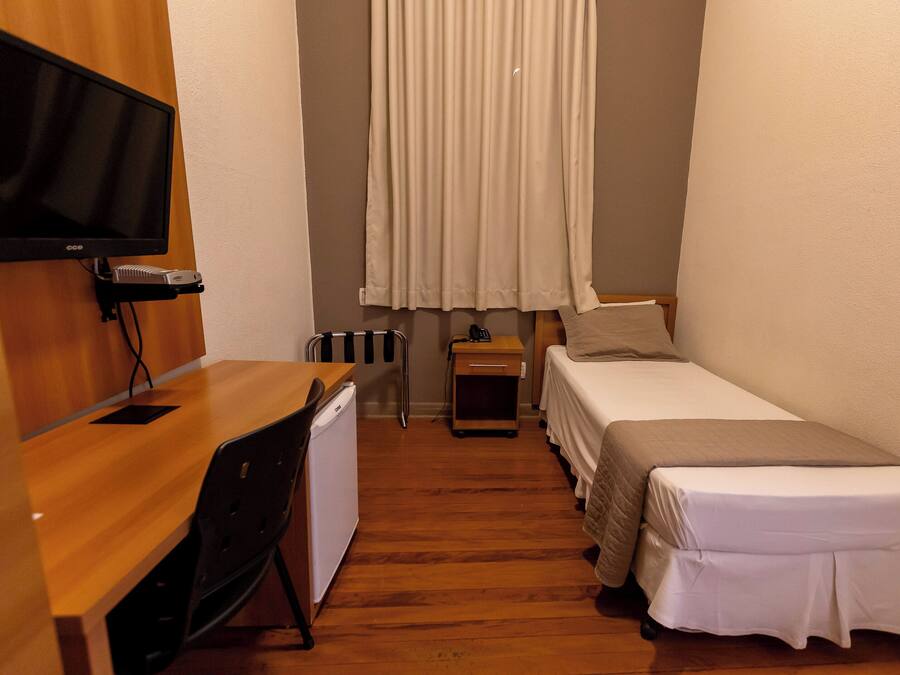 Una habitación pequeña y acogedora con escritorio de madera, cama, televisor montado en la pared y ventana con cortinas.