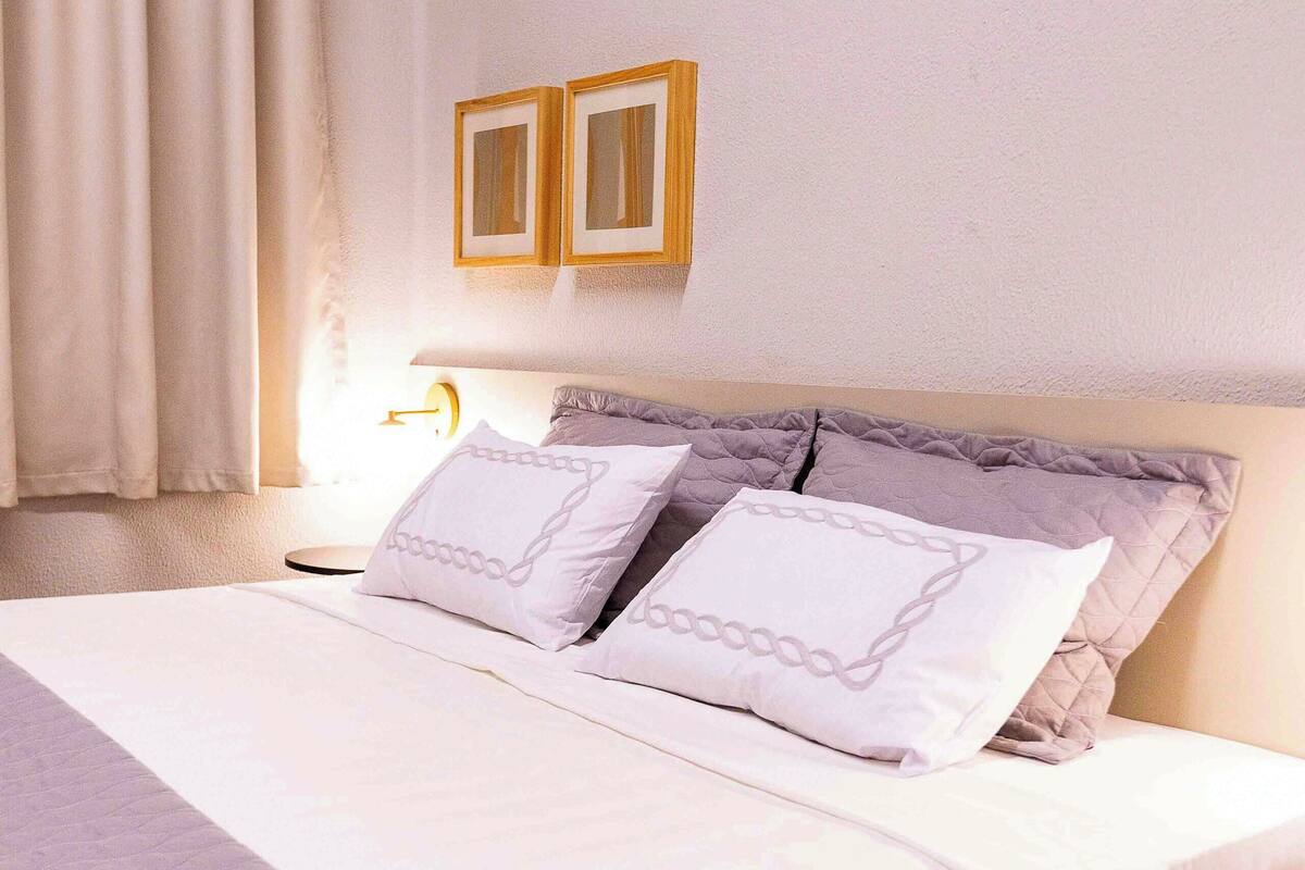 A neatly made bed with a quilted cover and two pillows, a bedside table with a lamp, and a wall-mounted picture frame.