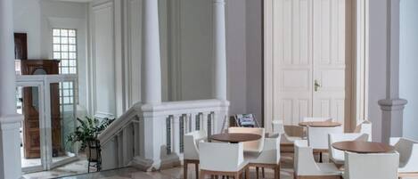 A spacious lobby with a grand staircase, white columns, and a seating area with round tables and chairs.