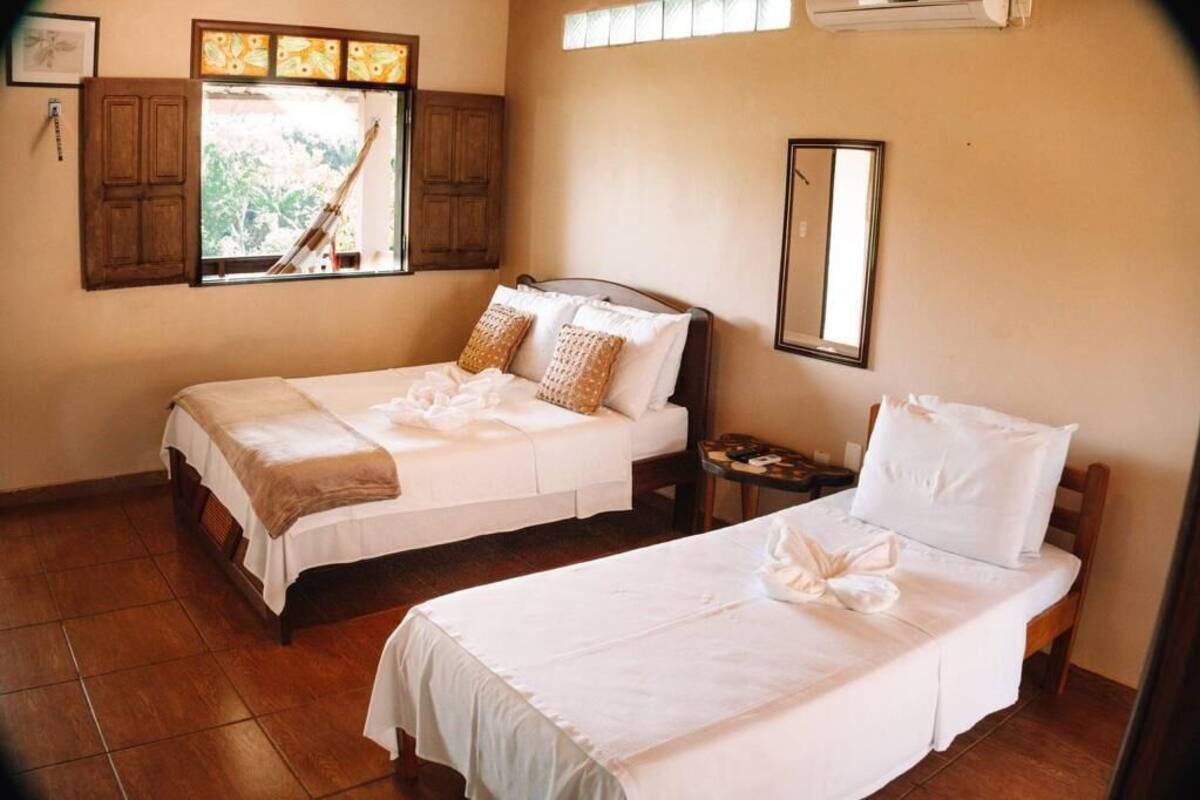 Une chambre d’hôtel avec deux lits simples, une armoire en bois, une fenêtre avec des rideaux, un miroir et une petite table.