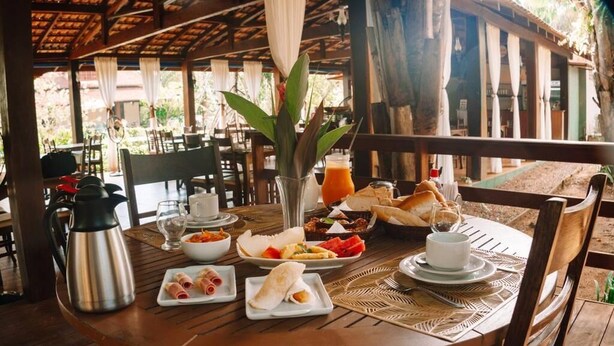 A wooden table set with breakfast items, including bread, fruit, and a beverage, in an outdoor dining area with a wooden ceiling and natural light. - Amazônia ParkSuítes (Novo Airão)